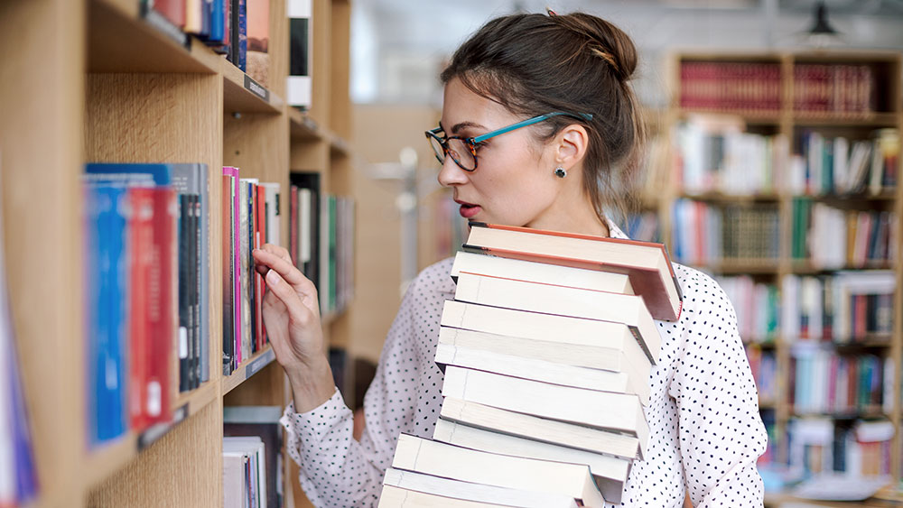 Eine junge Frau steht in einer Bibliothek vor einem Regal und hält einen Stapel Bücher in der Hand.