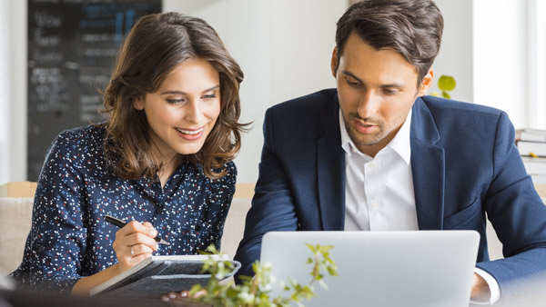 Eine Frau mit Stift und Notizblock und ein Mann sitzen an einem Laptop. Eine Frau mit Stift und Notizblock und ein Mann sitzen an einem Laptop.
