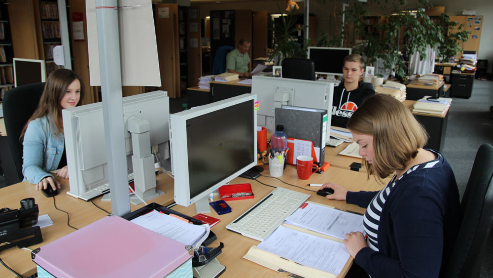 Drei Azubis sitzen an ihrem Schreibtisch in einem Großraumbüro vor ihrem PC und arbeiten.  Drei Azubis sitzen an ihrem Schreibtisch in einem Großraumbüro vor ihrem PC und arbeiten.
