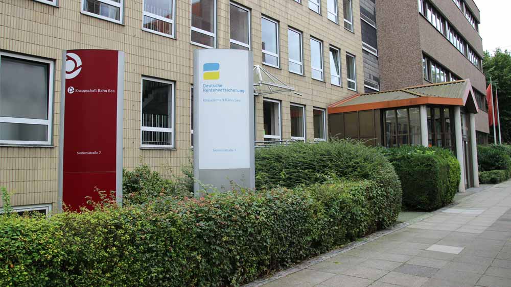 Vor der dem Bürogebäude der Regionaldirektion in Hannover stehen zwei hohe Stelen mit dem Logo der Knappschaft-Bahn-See und dem Logo der Deutschen Rentenversicherung. Eine grüne Hecke umgibt das Haus.