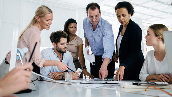 Sieben Mitarbeiterinnen und Mitarbeiter sitzen bzw. stehen an einem Tisch und sehen sich Unterlagen an.  Sieben Mitarbeiterinnen und Mitarbeiter sitzen bzw. stehen an einem Tisch und sehen sich Unterlagen an.