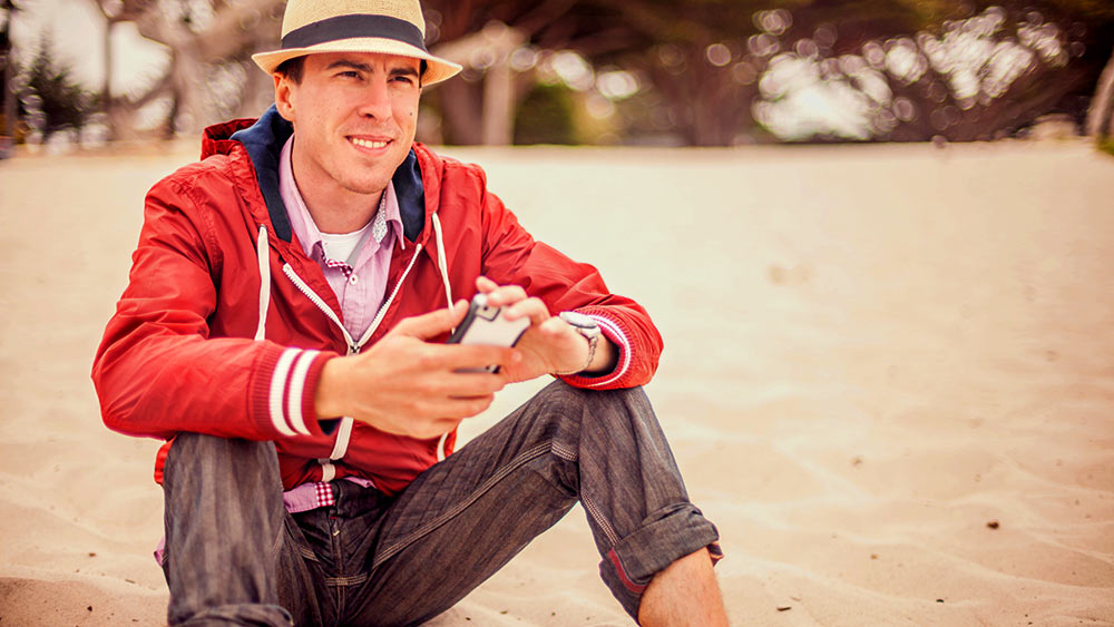 Ein Mann sitzt mit einem Smartphone in der Hand am Strand und schaut in die Ferne.