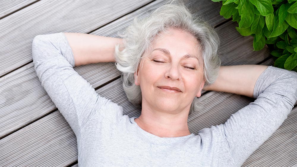 Eine Frau liegt entspannt mit geschlossenen Augen auf dem Holzboden einer Terrasse. Nahaufnahme des Gesichts.