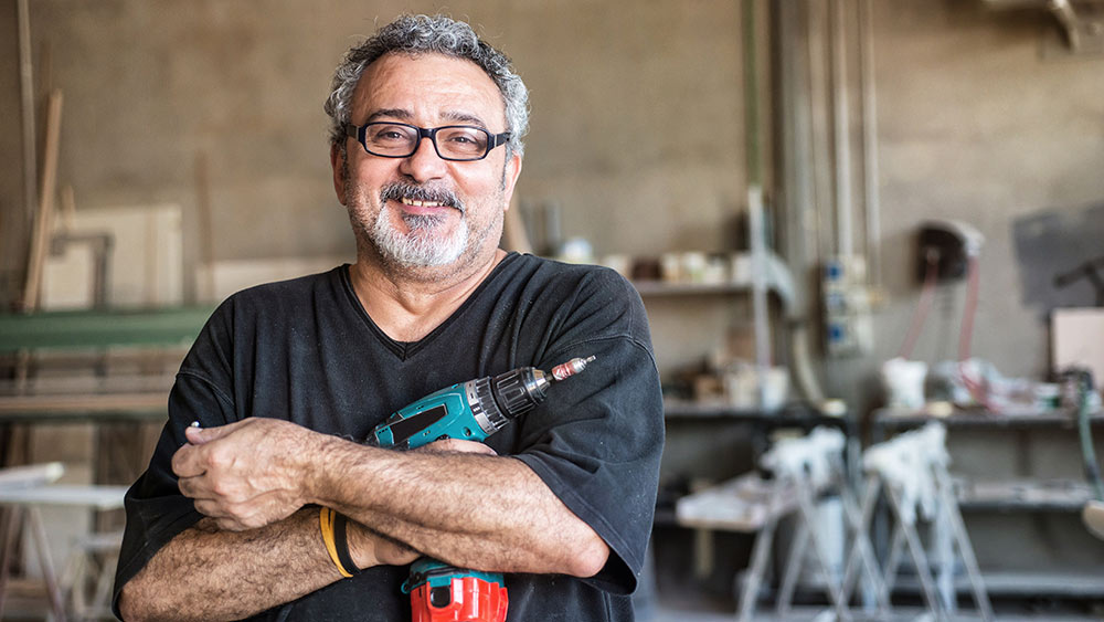 Ein Mann steht mit einer Bohrmaschiene in der Hand in einer Werkstatt.