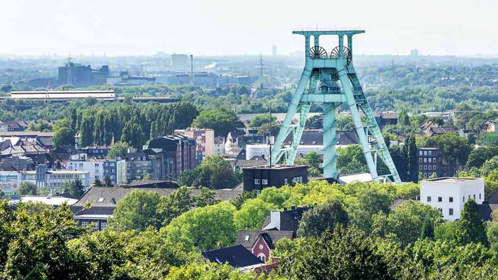 Inmitten einer städtischen Siedlung mit vielen grünen Bäumen steht der Förderturm einer Zeche. 