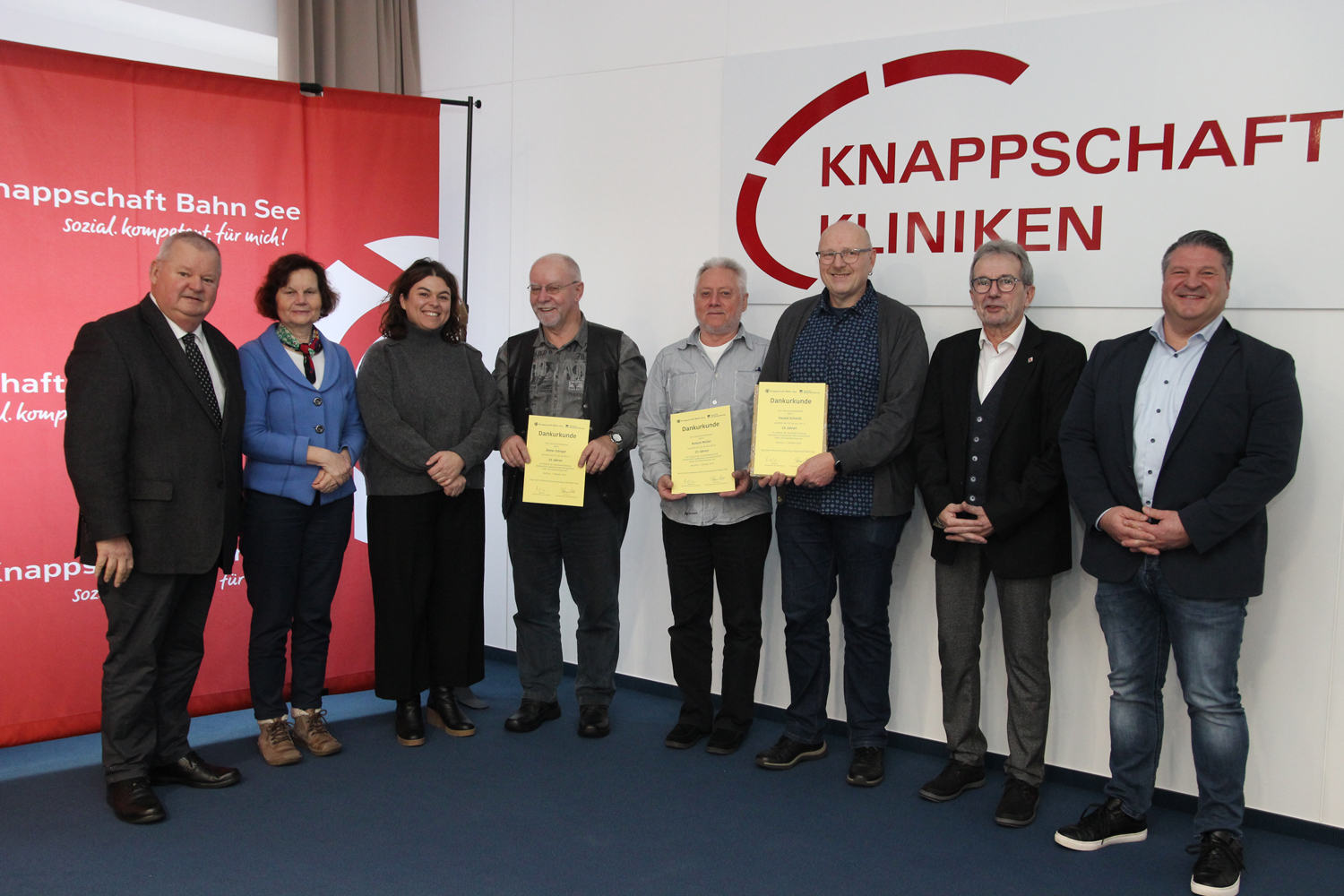 Versichertenberater der KBS und der Regionalausschuss der KBS im Saarland vor einer roten Rückwand mit KBS-Logo sowie einer Wand mit der Aufschrift Knappschaft Kliniken Versichertenberater der KBS und der Regionalausschuss der KBS im Saarland vor einer roten Rückwand mit KBS-Logo sowie einer Wand mit der Aufschrift Knappschaft Kliniken