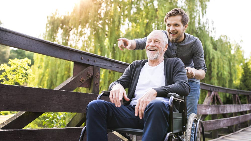 Ein junger Mann schiebt einen älteren Mann im Rollstuhl über eine Holzbrücke.   Ein junger Mann schiebt einen älteren Mann im Rollstuhl über eine Holzbrücke.