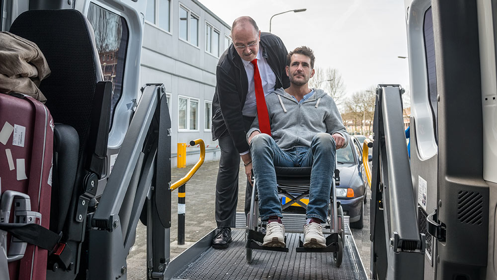 Ein Mann schiebt einen anderen Mann, der im Rollstuhl sitzt, in einen Krankentransportwagen.